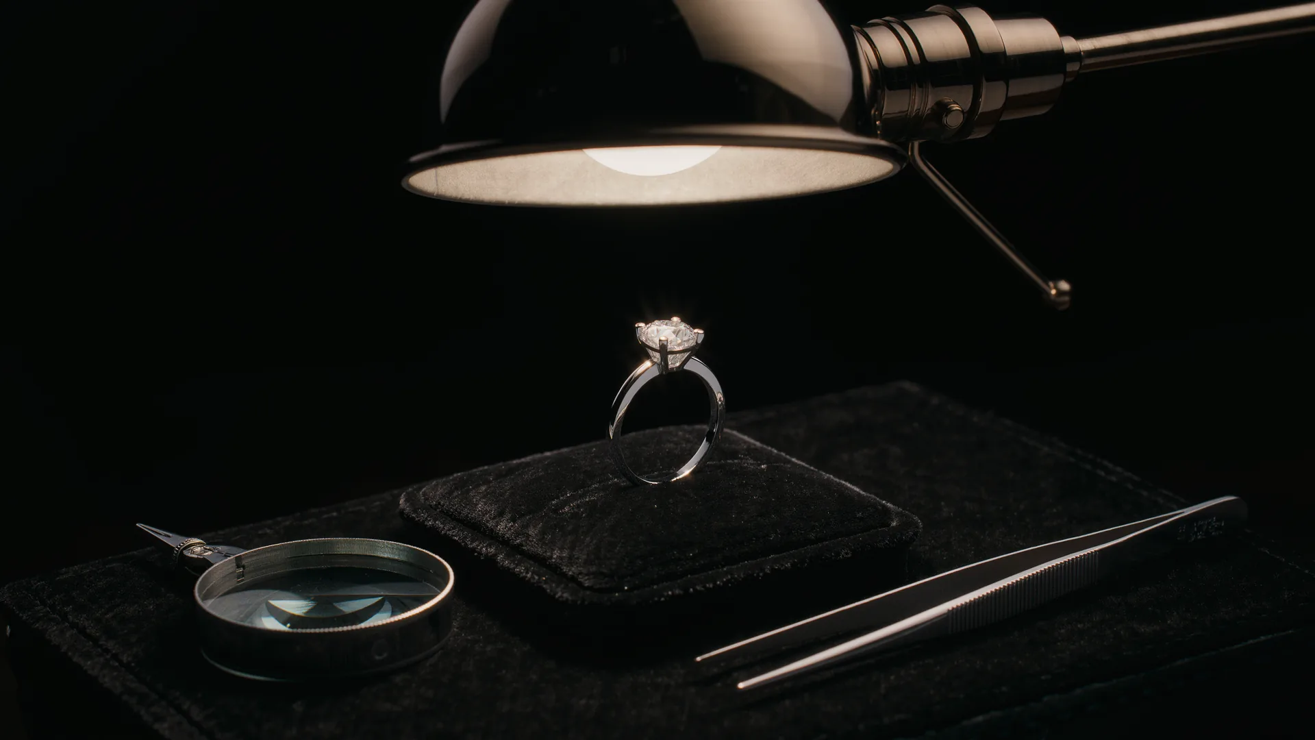 Anillo de diamantes sobre terciopelo negro bajo la lámpara del joyero, con lupa y pinzas