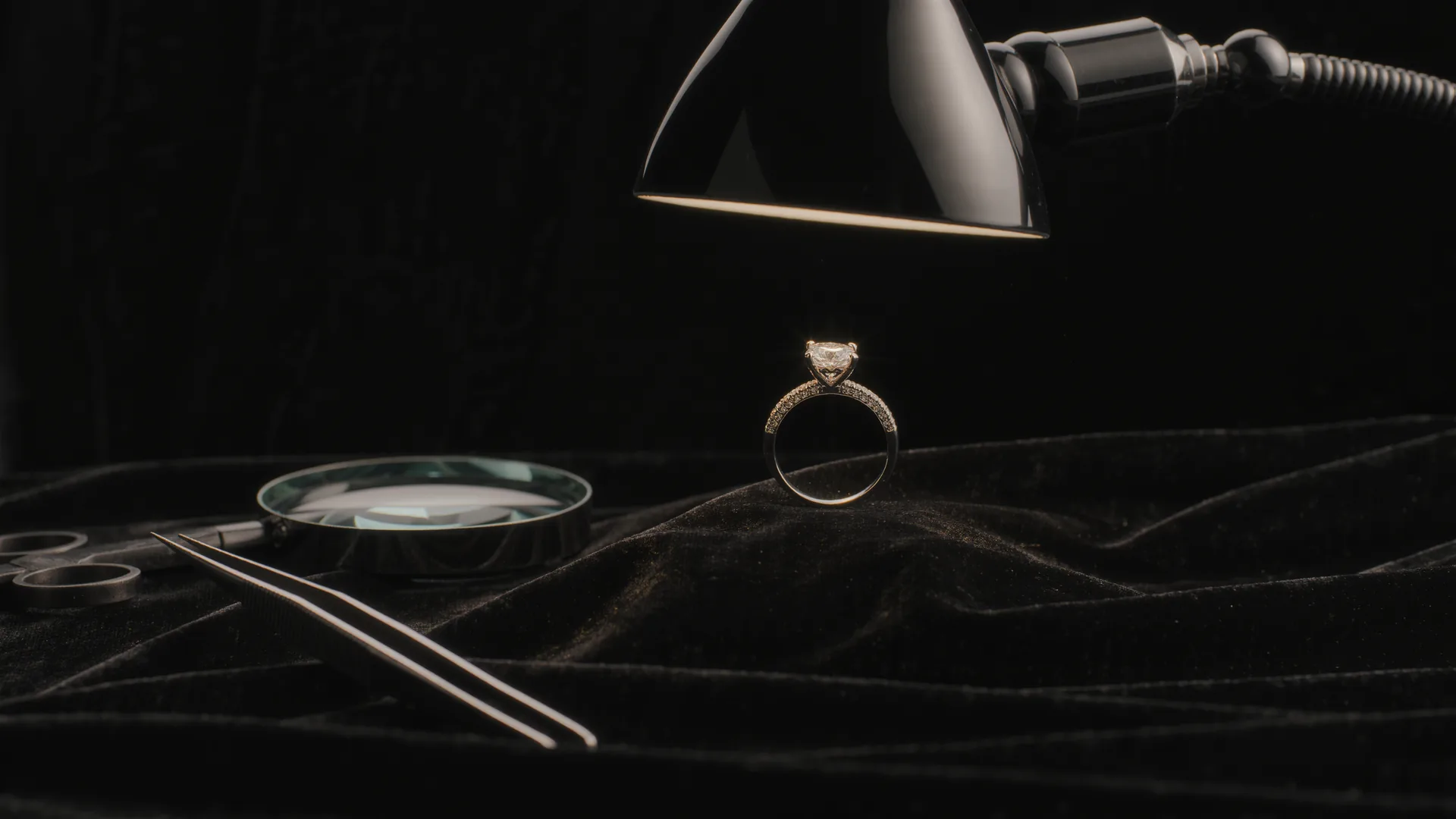 Anillo de diamantes sobre terciopelo negro bajo la lámpara del joyero, con lupa y pinzas